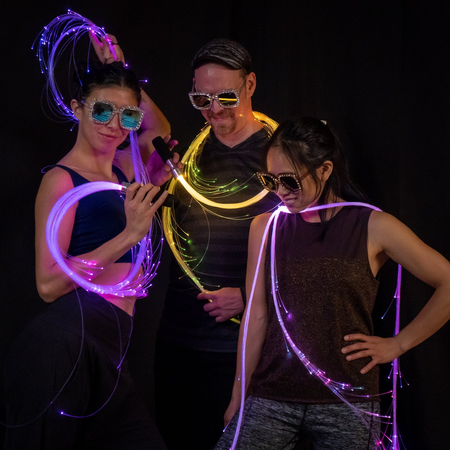 Bright led whips surround three people standing in a cool group pose, wearing reflective glasses. They look like they are in a photoshoot at a rave.